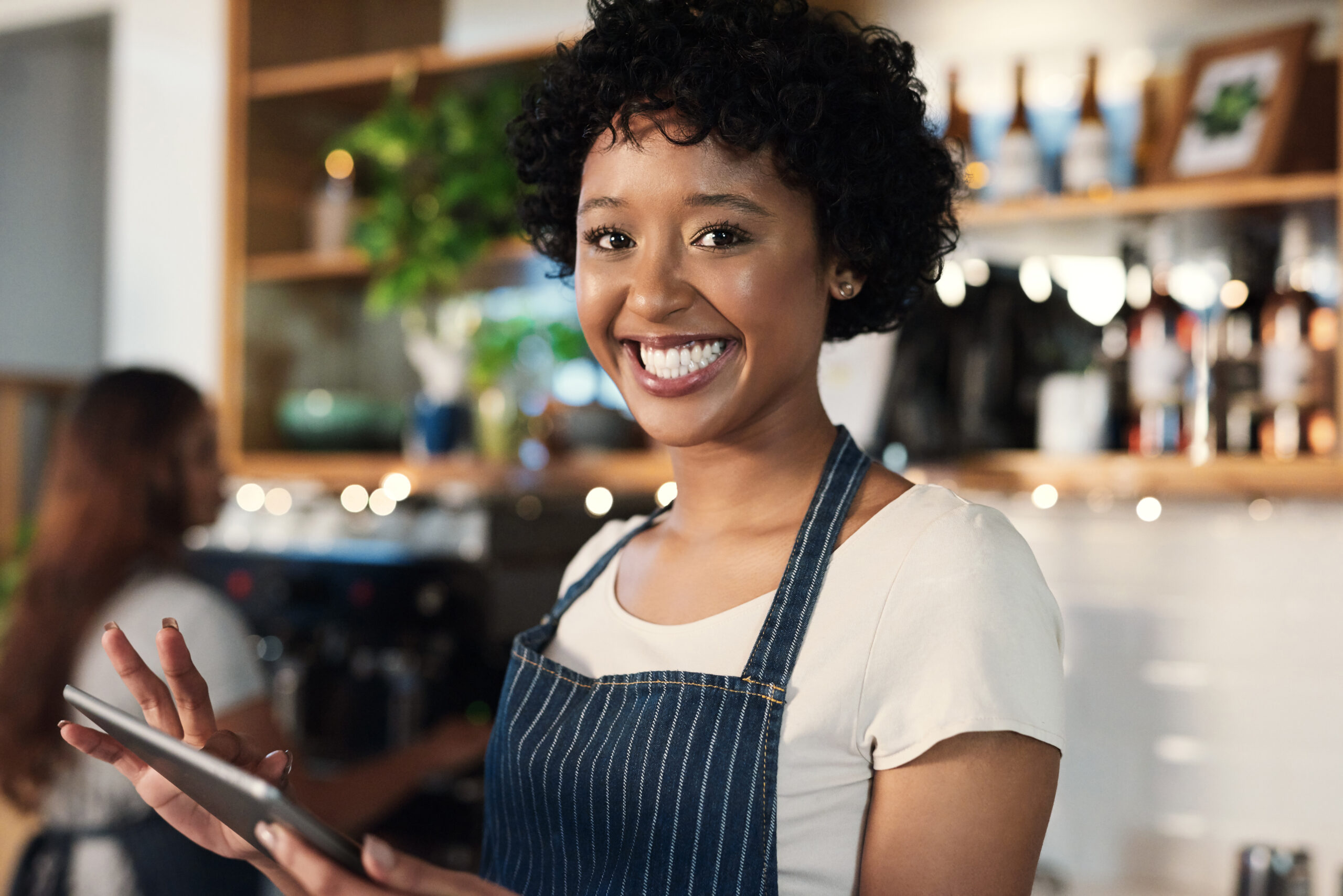 A woman in an apron efficiently manages orders on a tablet using inventory management software in a bustling restaurant.